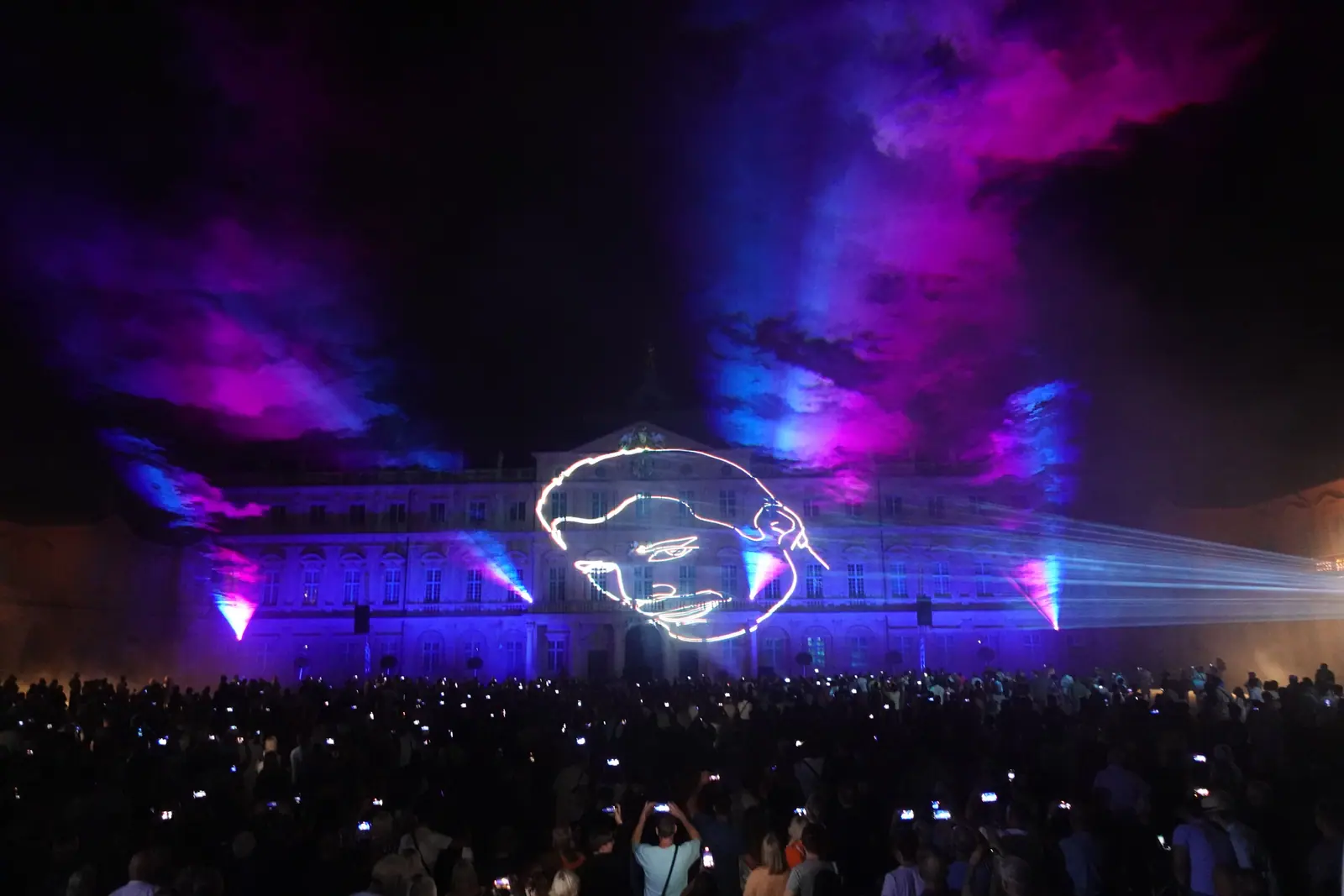 Lasershow vor dem Residenzschloss Rastatt: eine Linienzeichnung aus Laserlicht auf der Fassade, violette Wolken am Himmel, davor tausende Zuschauer mit erhobenen Handys